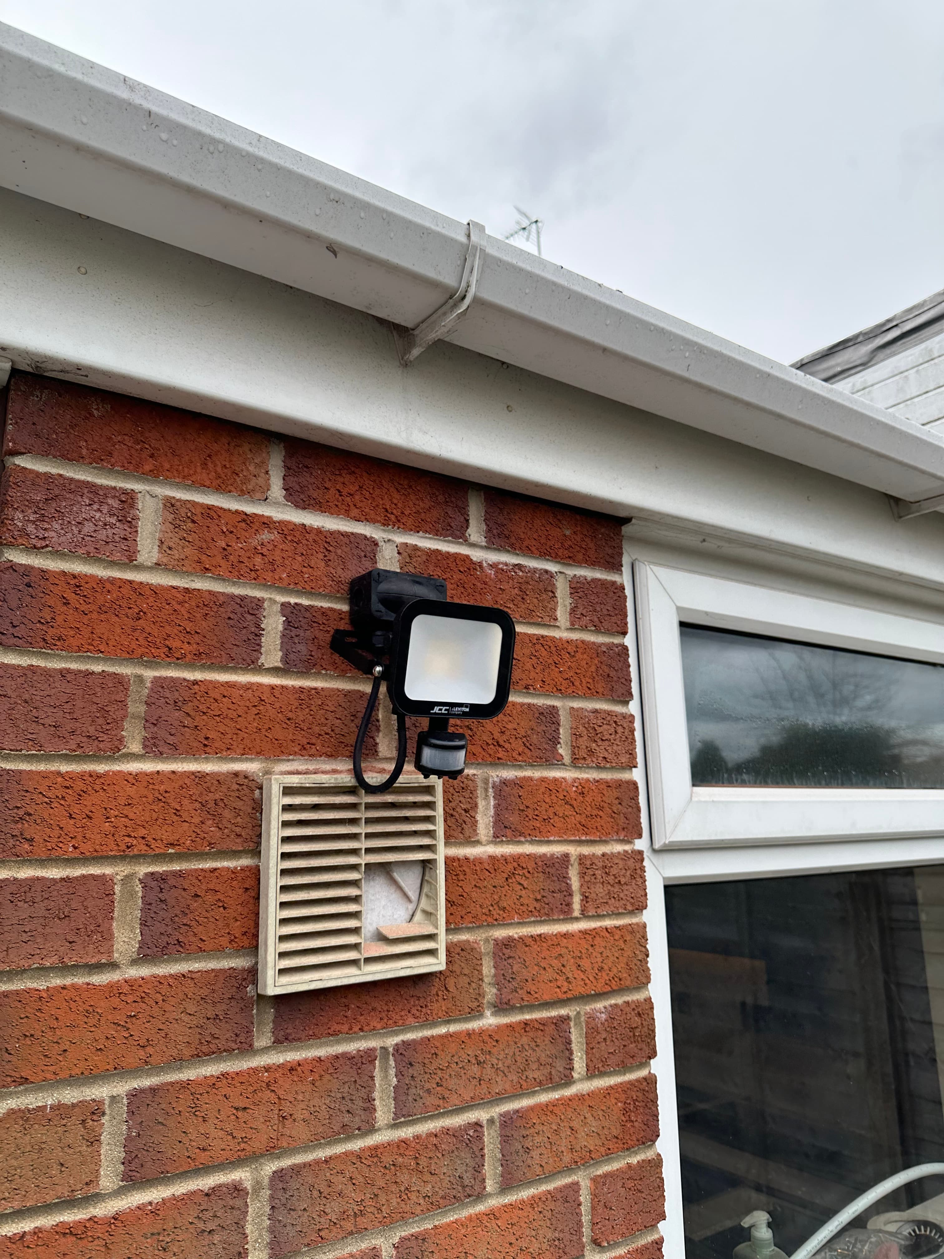 Black motion-sensor security light and beige air vent mounted on an exterior brick wall.