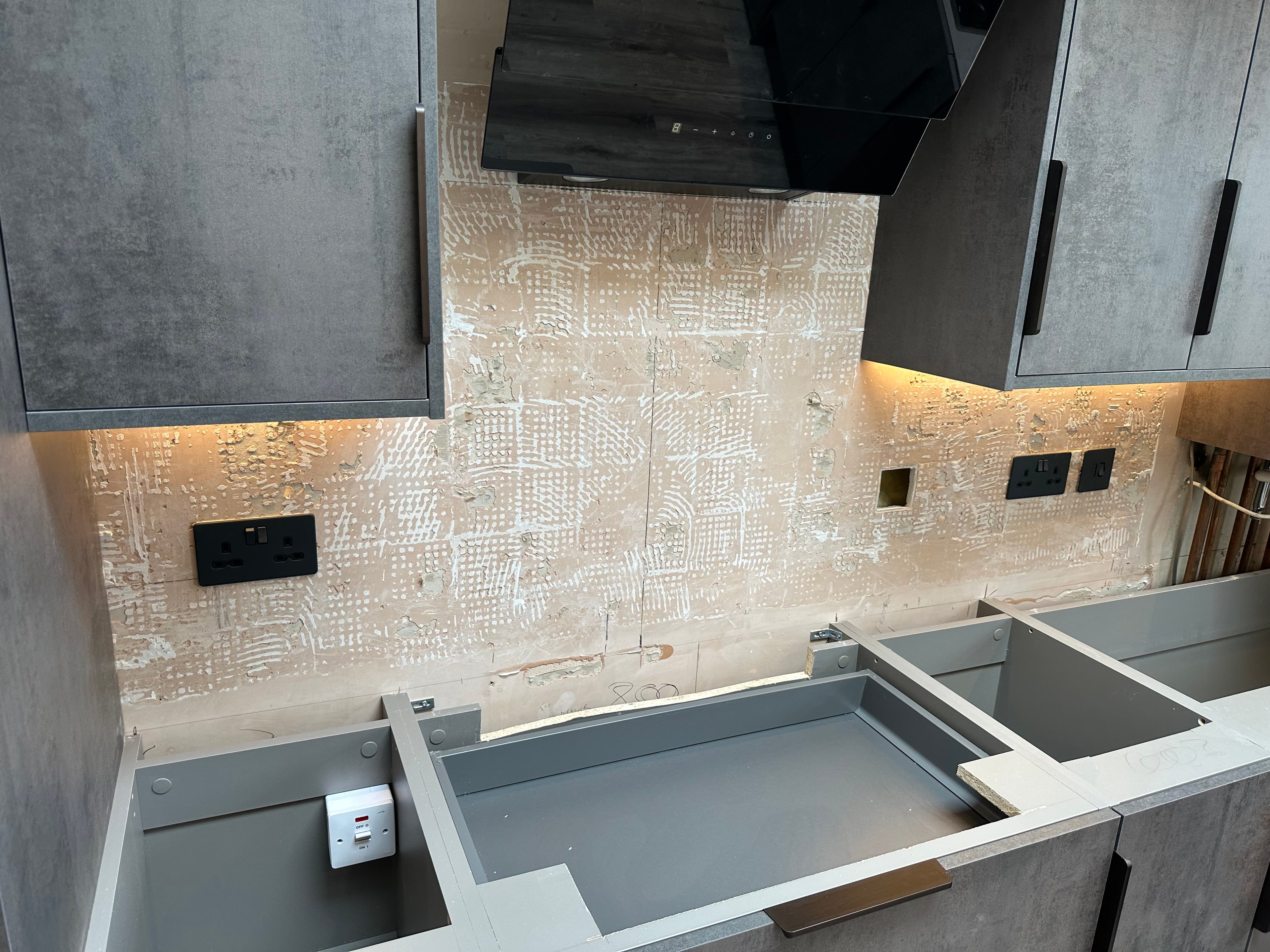 Modern grey kitchen cabinets during renovation with an unfinished backsplash and black extractor hood.